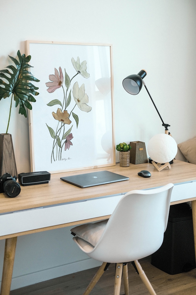 May include: A light wood desk with a white drawer, a laptop, a black speaker, a camera, a framed watercolor print of flowers, a lamp with a white moon-shaped lampshade, and a white chair with a light brown cushion.