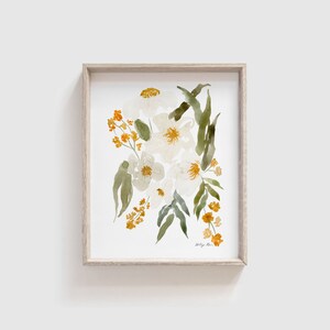 May include: Watercolor floral art print in a light wood frame. The artwork features white flowers with yellow centers, accented by green leaves and small orange blossoms. The artist's signature is visible in the lower right corner.