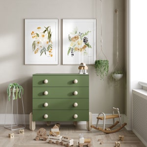 May include: A green dresser with four drawers and white knobs, flanked by framed floral art prints. A wooden rocking horse and toy train sit on the floor, creating a nursery or children's room theme. Hanging planters add a touch of greenery.