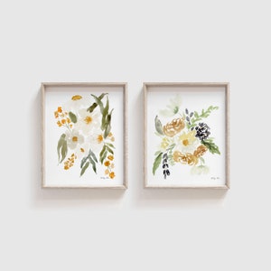 May include: Two framed watercolor floral prints. One features white flowers with orange accents and green leaves. The other print showcases yellow, gold, and dark blue flowers with green foliage. Both are in light wood frames.