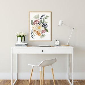 May include: A framed watercolor print of a floral bouquet with a white background. The flowers are in shades of pink, yellow, orange, and blue. The print is hanging on a white wall above a white desk with a white chair.