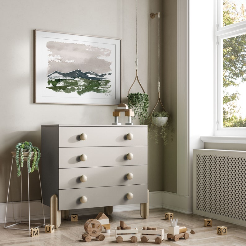 May include: A light gray four-drawer dresser with wooden knobs and legs. A framed watercolor painting of mountains hangs above the dresser. Wooden toy train and blocks are on the floor. Hanging planters and a metal plant stand add greenery to the room.