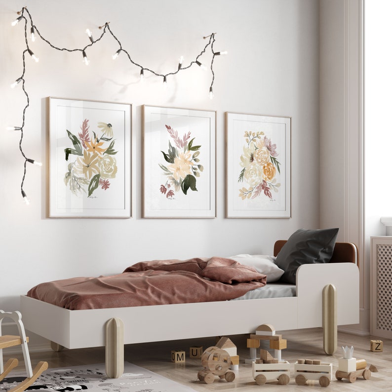 May include: Three framed watercolor prints of floral bouquets in shades of yellow, pink, and green. The prints are hanging on a white wall above a white bed with a pink blanket.