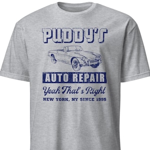 May include: A grey t-shirt with a blue graphic. The design features the text "PUDDY'S AUTO REPAIR" above a car illustration. Below the car, the text reads "Yeah That's Right" and "NEW YORK, NY SINCE 1995".