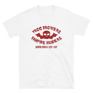 May include: White t-shirt with a red vintage-style logo that says "Frog Brothers Vampire Hunters Santa Carla Est. 1987". The logo features a skull with bat wings.