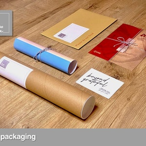 May include: A brown cardboard tube with a white label that says "The Minimalist Vibe" is used to package a rolled poster. The poster is wrapped in a blue and white paper with a string tied around it. A red envelope with a white string tied around it and a white card with the text "beyond grateful" are also included in the packaging.