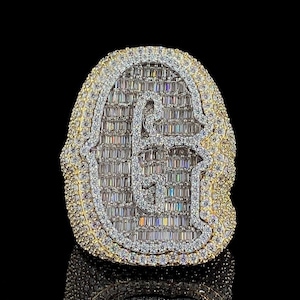 Pode incluir: Um anel de ouro com uma grande letra 'G' cravejada em pavé no centro. O anel é rodeado por diamantes redondos menores.