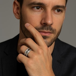 Può includere: Un anello d'argento con un design rettangolare e una fascia di piccole pietre trasparenti. L'anello è al dito di una persona, che indossa una giacca scura. Lo sfondo è grigio.