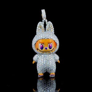 Puede incluir: Un colgante plateado con forma de personaje de dibujos animados con un disfraz de conejito, incrustado con piedras brillantes. El personaje tiene una cara naranja, ojos morados y una expresión sonriente. El colgante tiene una argolla.