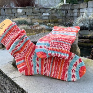 May include: A pair of colorful knitted socks with a matching cuff, featuring a pattern of red, orange, and blue stripes and diamond shapes. The socks are displayed on a wooden form with the size 40/41.