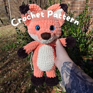 May include: A plush, crocheted fox toy in shades of peach, white, and brown. The fox has large blue eyes and the words "Crochet Pattern" are written across the top. The fox is being held up by a person.