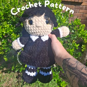 May include: Crochet pattern for a black and white doll with a white face and black hair. The doll is wearing a black dress with a white collar and white cuffs. The doll is holding a white crocheted flower.