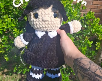 May include: Crochet pattern for a black and white doll with a white face and black hair. The doll is wearing a black dress with a white collar and white cuffs. The doll is holding a white crocheted flower.