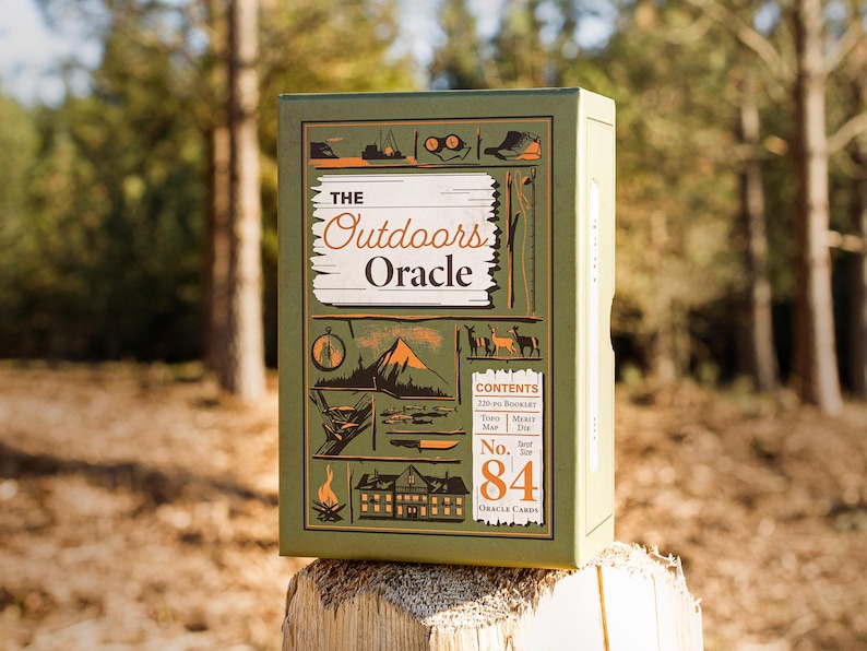 A forest green oracle deck box titled "The Outdoors Oracle" sits on a rustic trail fence post, flanked by a warm pine forest setting. Black and copper foil illustrations of mountains, deer, and other outdoors scene can be seen crisply on the box.