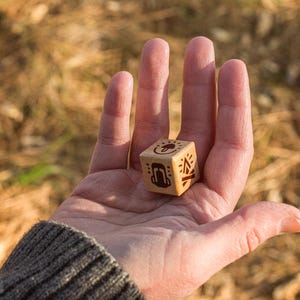 A close up shot of a women's hand is seen holding a wooden, six sided, merit die with a warm forest floor in the background. Three sides of the die are seen with a mushroom, backpack, and fire symbol in view. Warm sunlight hits from the right.