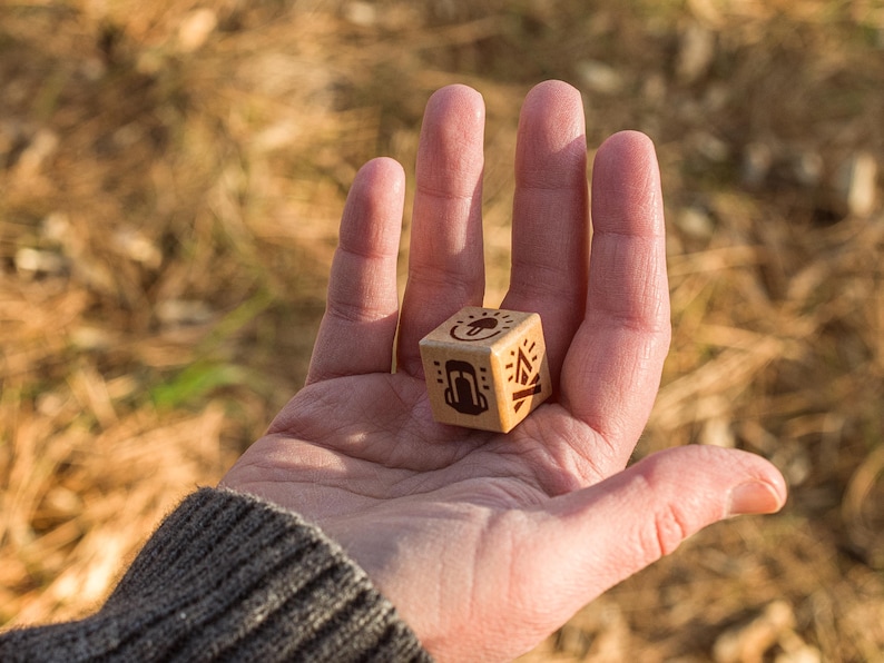 A close up shot of a women's hand is seen holding a wooden, six sided, merit die with a warm forest floor in the background. Three sides of the die are seen with a mushroom, backpack, and fire symbol in view. Warm sunlight hits from the right.