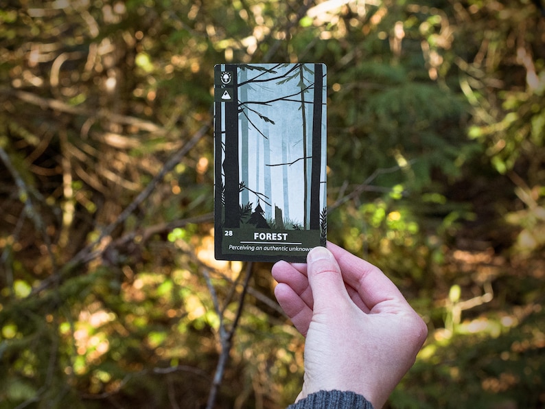 An oracle card titled "Forest" being held outside with trees and foliage behind it. Flavor text on the card reads "Perceiving an authentic unknown". A mushroom and mountain symbol along with the number 28 can be seen on the card.