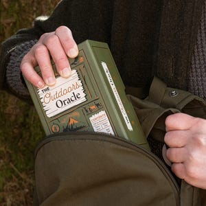 The Outdoors Oracle box is seen being taking out of a green linen backpack with a tree in the background.
