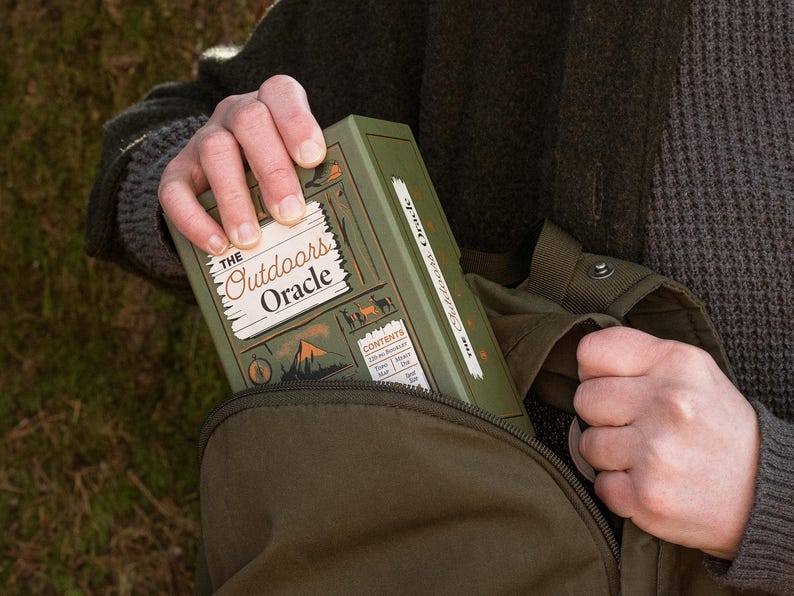 The Outdoors Oracle box is seen being taking out of a green linen backpack with a tree in the background.