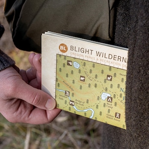 A close up shot of a folded topographical map being taken out of a coat pocket. The words "Blight Wilderness" and "Oregon Parks" can be seen, as well as some park symbols, dotted trails, and water symbols.