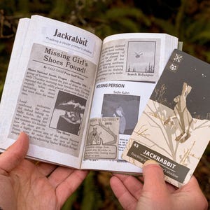 A hand holds open a book with the title "Jackrabbit" with several clippings from newspapers concerning a missing person. A card is held next the book, also titled "Jackrabbit" with an illustration of a rabbit jumping in a dark snowy setting.
