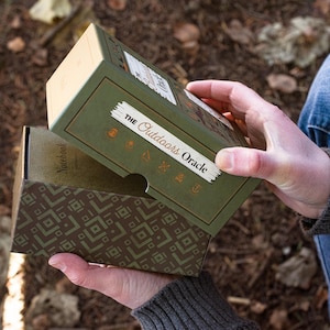 The Outdoors Oracle is seen being opened next to a forest floor setting. Six copper symbols can be seen on the the top of the box, with an interesting retro outdoor pattern being reveled as the bottom lip of the box is exposed.