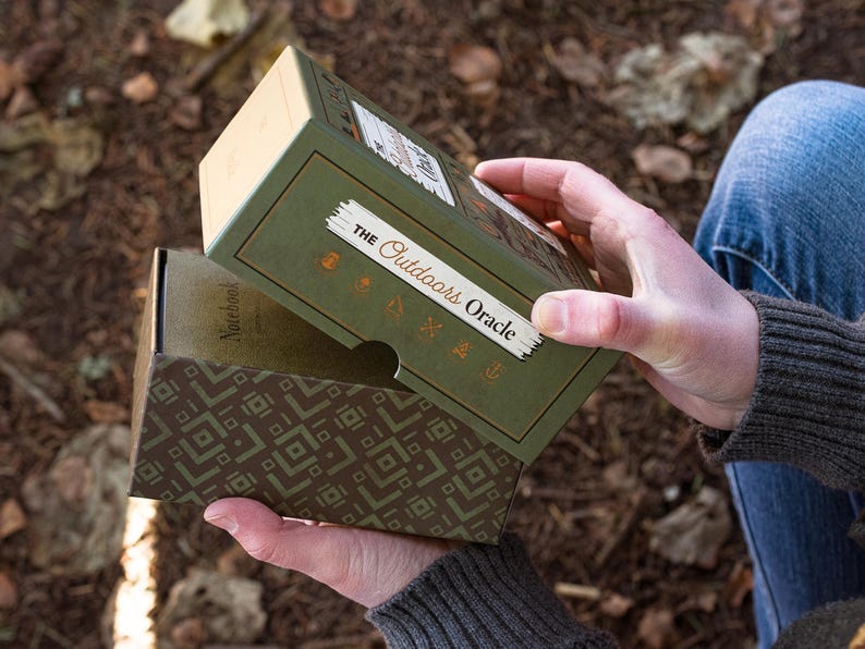 The Outdoors Oracle is seen being opened next to a forest floor setting. Six copper symbols can be seen on the the top of the box, with an interesting retro outdoor pattern being reveled as the bottom lip of the box is exposed.