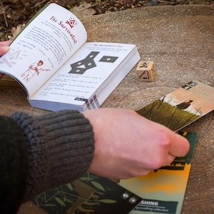 A hand is seen laying down an oracle card on a large wooden stump, completing the last card in a spread.  The second hand holds open a booklet showing illustrated details about how the card spread is performed. A wooden six sided die is seen as well.