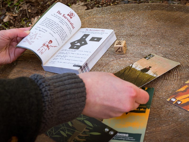 A hand is seen laying down an oracle card on a large wooden stump, completing the last card in a spread.  The second hand holds open a booklet showing illustrated details about how the card spread is performed. A wooden six sided die is seen as well.