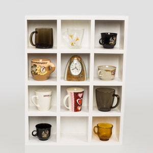 Può includere: Un'unità a scaffale bianca con 12 scomparti, ognuno contenente una diversa tazza in ceramica o vetro. Le tazze sono di vari colori e motivi, tra cui disegni floreali, geometrici e astratti.