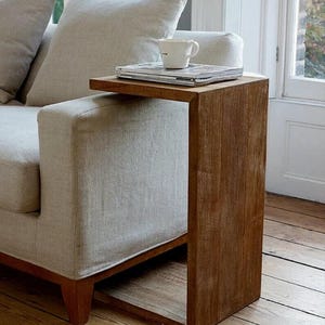 Op de afbeelding: Een houten bijzettafel met een warme bruine afwerking staat naast een lichtgekleurde bank. Een witte mok en een stapel tijdschriften liggen op het tafelblad. De tafel heeft een eenvoudig, rechthoekig ontwerp en is ongeveer 60 cm hoog.
