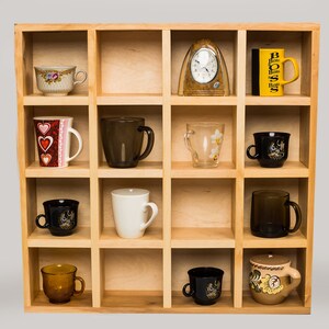 Può includere: Un'unità a mensola in legno con 12 scomparti, ognuno contenente una tazza o una tazza diversa. Le tazze sono di vari colori e motivi, tra cui nero, bianco, marrone, rosso e giallo. C'è anche un orologio e un libro sullo scaffale.