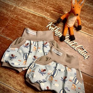 Könnte beinhalten: Zwei Paar Baby-Blümchen mit einem blauen und weißen Druck von Heißluftballons, Wolken und Eichhörnchen. Die Blümchen haben einen braunen Bund und eine Tasche auf jeder Seite.