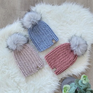 May include: Three knitted beanies in pastel colors: light pink, periwinkle blue, and rose. Each hat features a large, fluffy, gray faux fur pom-pom on top. The beanies are displayed on a white, fluffy rug.