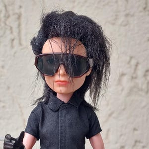 May include: A doll with black hair and sunglasses, wearing a black short-sleeved shirt, black pants, and gloves. The doll is holding a cigarette. The doll has a belt and a bracelet with studs.