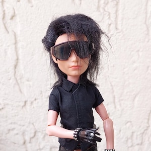May include: A doll dressed in a black short-sleeved shirt, black pants, and sunglasses. The doll has black hair and is wearing black gloves and studded bracelets. The doll is standing on a metal table.