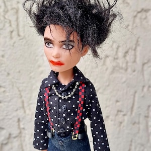 Könnte beinhalten: Eine Puppe mit zerzaustem schwarzem Haar, dunklem Augen-Make-up und leuchtend rotem Lippenstift. Die Puppe trägt ein schwarzes langärmeliges Hemd mit weißen Punkten, rot-schwarz karierte Hosenträger, eine silberne Kugelkette und eine blaue Jeans.