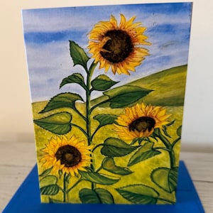 May include: A watercolor painting of sunflowers in a field. The painting features three sunflowers with yellow petals and dark brown centers, set against a backdrop of a green hill and a blue sky. The painting is on a rectangular card.