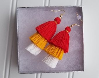 Puede incluir: Un par de pendientes de borla en capas con borlas blancas, amarillas y rojas. Los pendientes están en una pequeña caja de regalo.