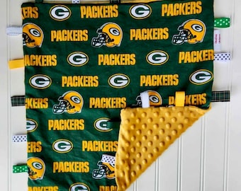 May include: Green and gold baby blanket with a green and white patterned fabric featuring the Green Bay Packers logo and the word "Packers" repeated throughout. The blanket has a yellow minky fabric on the back side and has multiple fabric tags attached to the edges.