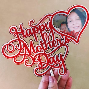 Puede incluir: Topper de pastel rojo y plateado con brillo con las palabras "Happy Mother's Day" en una fuente cursiva. Un marco en forma de corazón con una foto de una mujer y un niño está en la parte superior del topper.