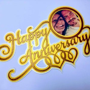 May include: Gold and white cake topper with the words "Happy Anniversary" and a photo of a couple in a circle.