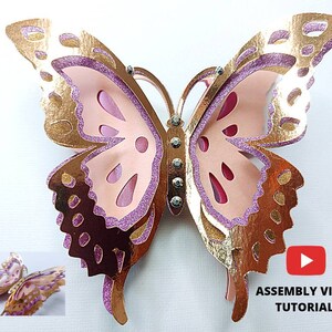 May include: A layered paper butterfly in shades of gold, pink, and purple. The butterfly has intricate cut-out details and is embellished with rhinestones. A smaller version of the butterfly is in the lower left corner. The text "ASSEMBLY VIDEO TUTORIAL" is in the lower right corner.