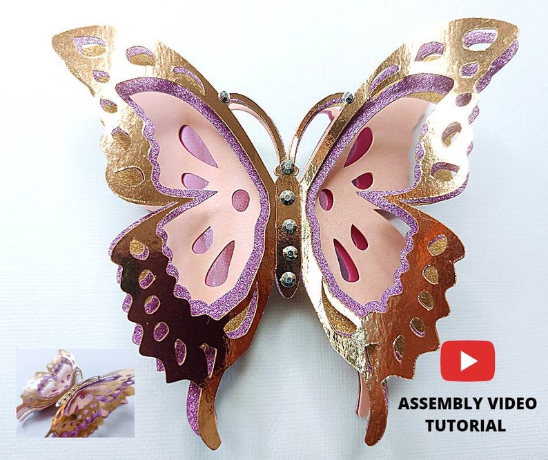 May include: A layered paper butterfly in shades of gold, pink, and purple. The butterfly has intricate cut-out details and is embellished with rhinestones. A smaller version of the butterfly is in the lower left corner. The text "ASSEMBLY VIDEO TUTORIAL" is in the lower right corner.