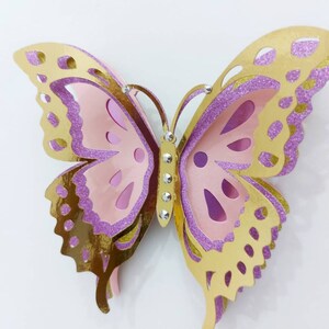 May include: A layered paper butterfly in shades of gold, pink, and purple. The butterfly has intricate cut-out details and is embellished with small, sparkling rhinestones. The wings have a glittery texture.
