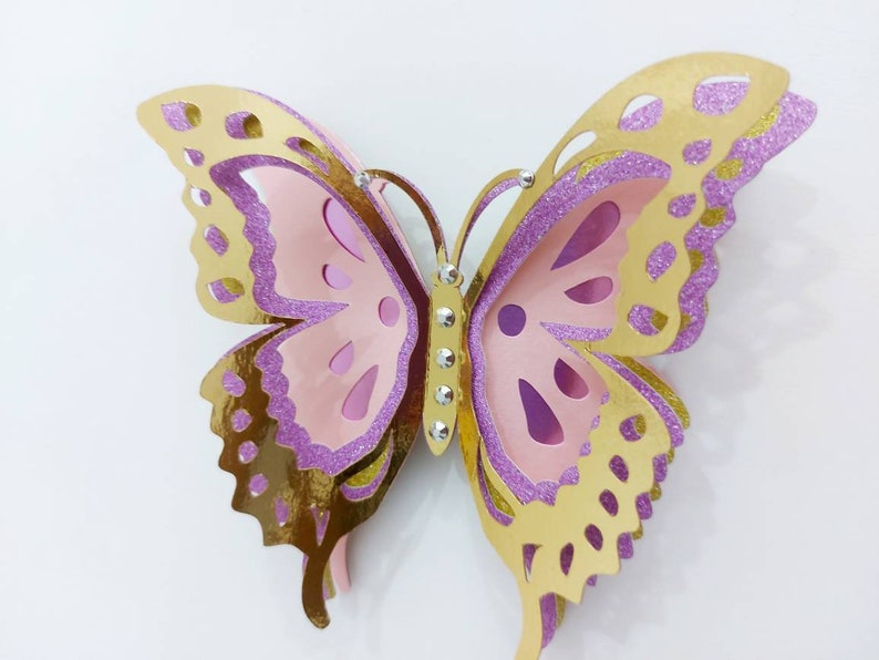 May include: A layered paper butterfly in shades of gold, pink, and purple. The butterfly has intricate cut-out details and is embellished with small, sparkling rhinestones. The wings have a glittery texture.