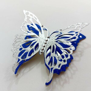 Peut inclure: Une d&eacute;coupe de papier en forme de papillon argent&eacute; et bleu avec des accents de paillettes. Le papillon a un design d&eacute;licat et complexe et est parfait pour ajouter une touche de fantaisie &agrave; tout projet d'artisanat.