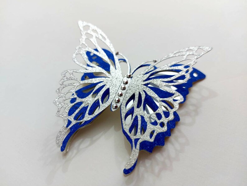 Peut inclure: Une d&eacute;coupe de papier en forme de papillon argent&eacute; et bleu avec des accents de paillettes. Le papillon a un design d&eacute;licat et complexe et est parfait pour ajouter une touche de fantaisie &agrave; tout projet d'artisanat.