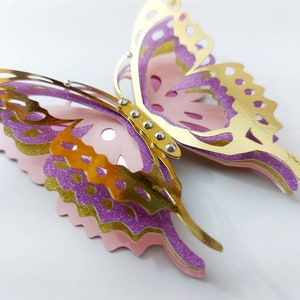 May include: A layered paper butterfly in gold, purple, and pink. The butterfly has intricate cut-out details on its wings and is embellished with small silver rhinestones. The wings are layered with glitter accents.