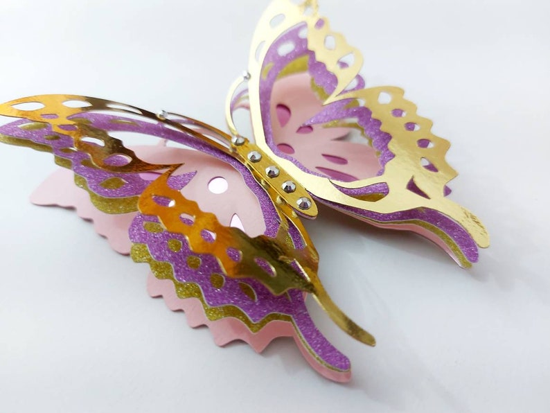 May include: A layered paper butterfly in gold, purple, and pink. The butterfly has intricate cut-out details on its wings and is embellished with small silver rhinestones. The wings are layered with glitter accents.